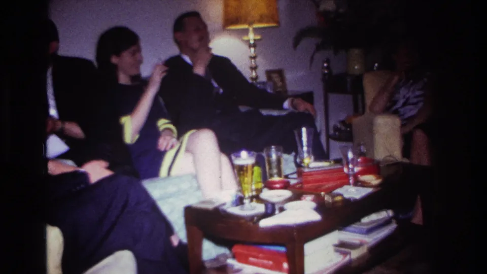 Adults in a 1967 Rockaway Beach living room appear to converse while seated near a coffee table with drinks, cigarettes, and books.