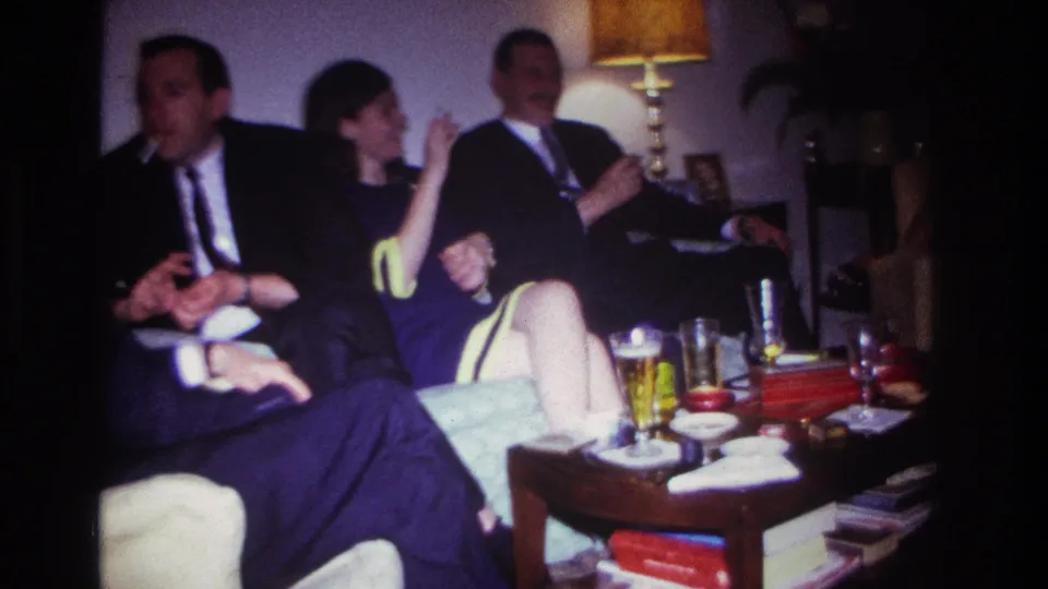 1967 Rockaway Beach home-movie frame with suited men and a woman seated close together around a living room coffee table.