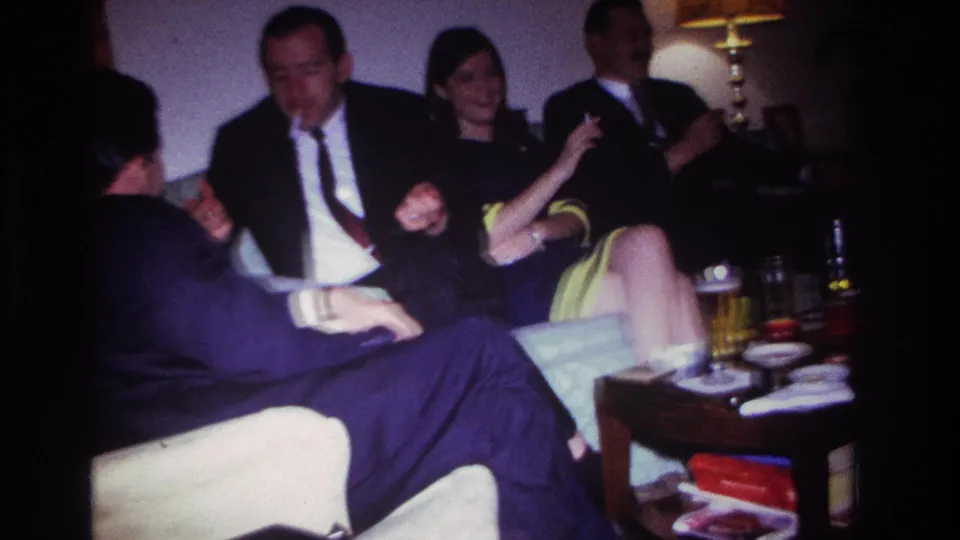 Adults in a private Rockaway Beach living room socialize around a low table with drinks, cigarettes, books, and a warm lamp nearby.