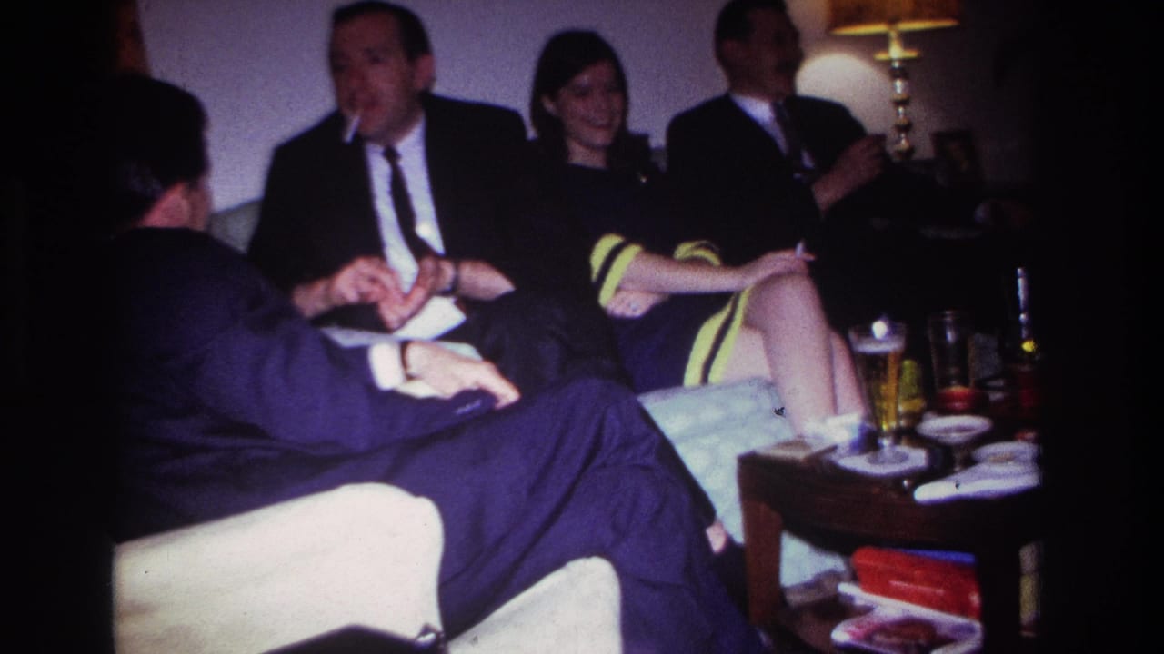 1967 Rockaway Beach home-movie frame showing adults in suits and a dark dress socializing in a dim private living room.