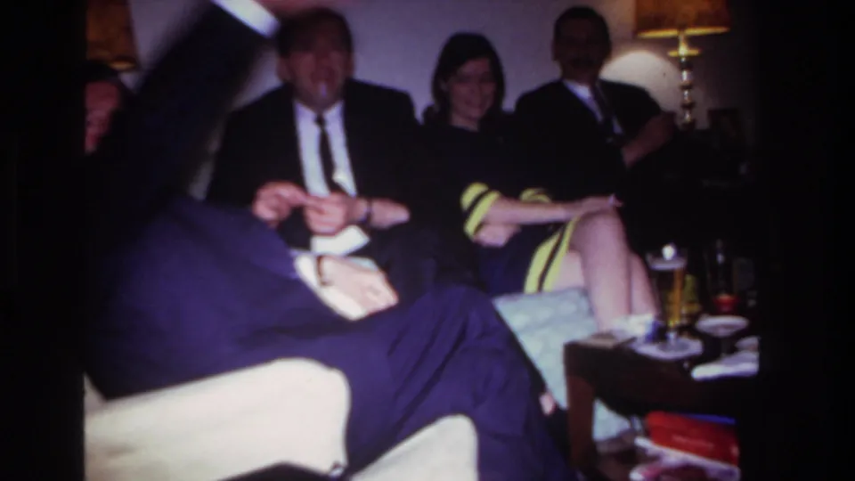 A handheld 8mm frame from the 1967 Rockaway Beach gathering, with motion blur crossing adults seated indoors.