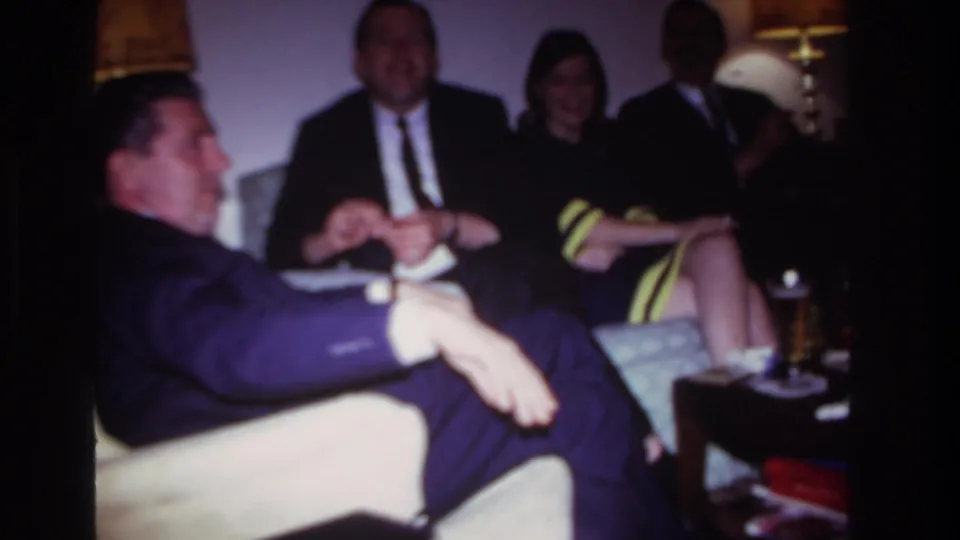 1967 Rockaway Beach home-movie frame showing a suited man in the foreground with friends seated behind him in a private living room.