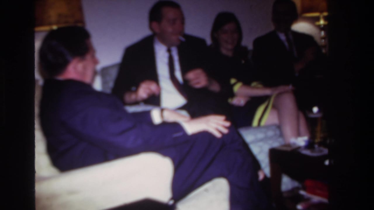 Adults in formal clothing sit in a living room with lamps and upholstered seating.