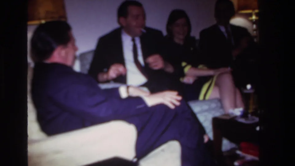 1967 Rockaway Beach home-movie frame showing adults in suits and dresses conversing in a dim living room.