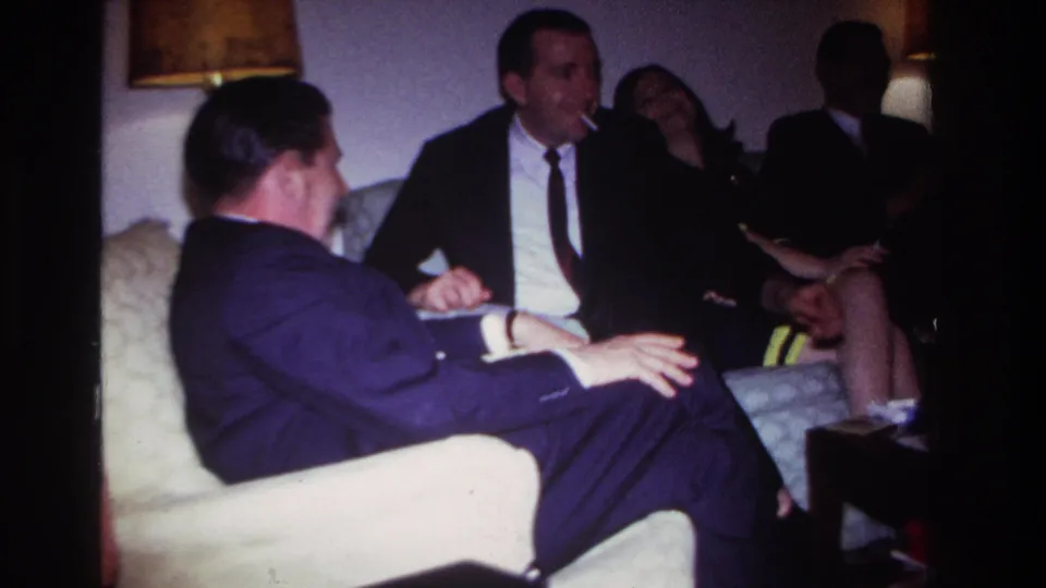 1967 Rockaway Beach interior frame with a suited man smoking while adults converse on sofa seating.