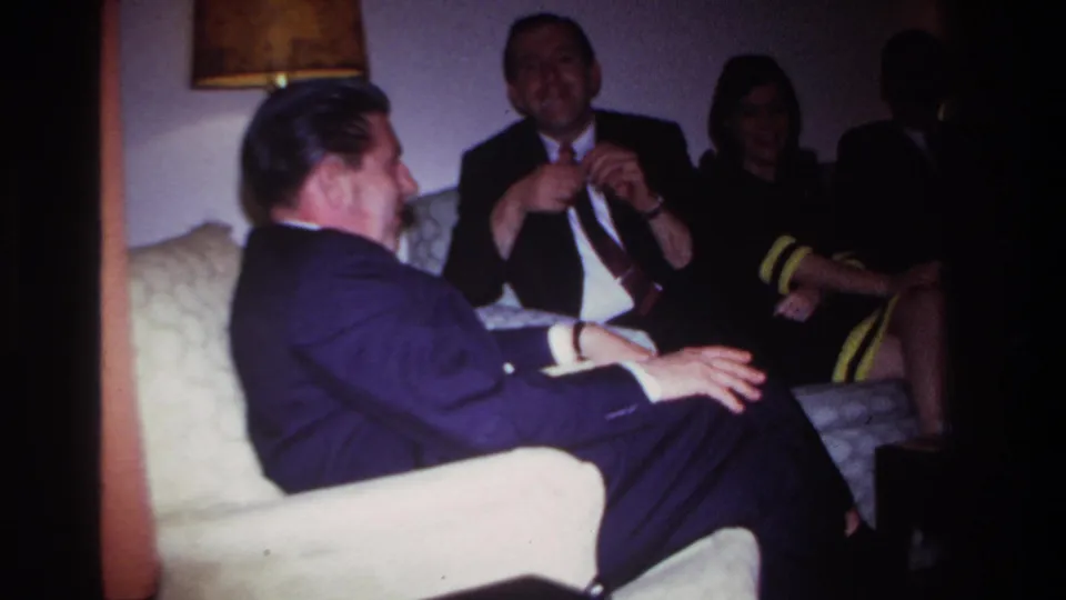1967 Rockaway Beach home-movie frame showing a seated group in formal attire during living-room conversation.