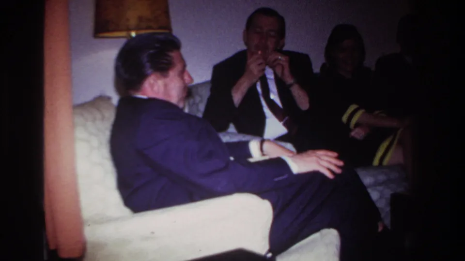 1967 Rockaway Beach living-room frame with adults in formal clothes talking as one man gestures near his face.