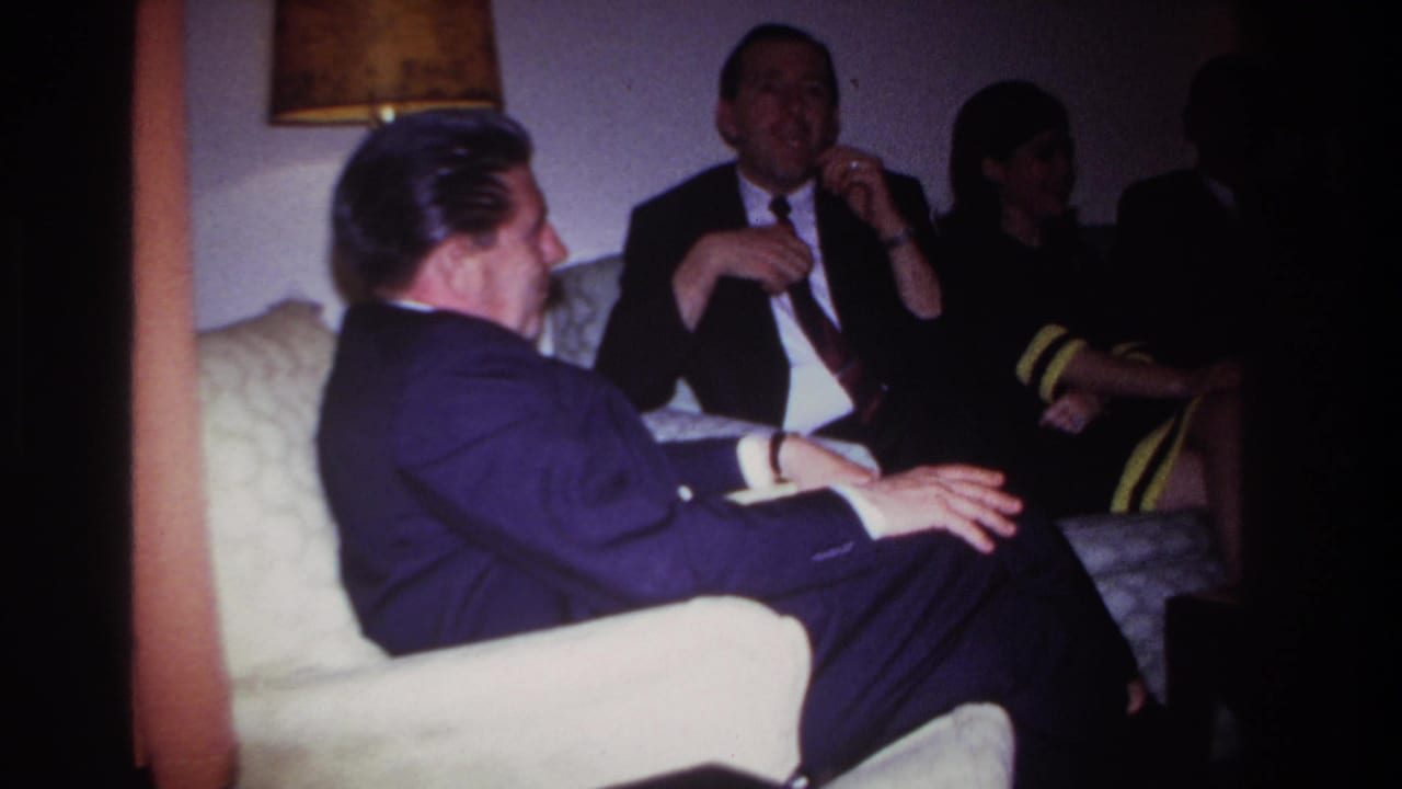 Adults in suits seated in a dim living room during an indoor gathering.