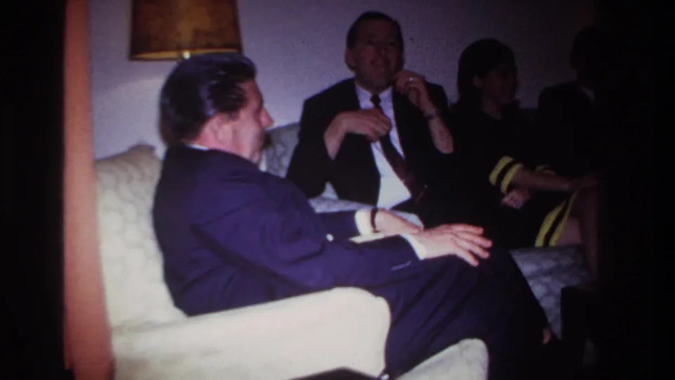 1967 Rockaway Beach home-movie frame showing adults in suits and a woman seated in a private living room.