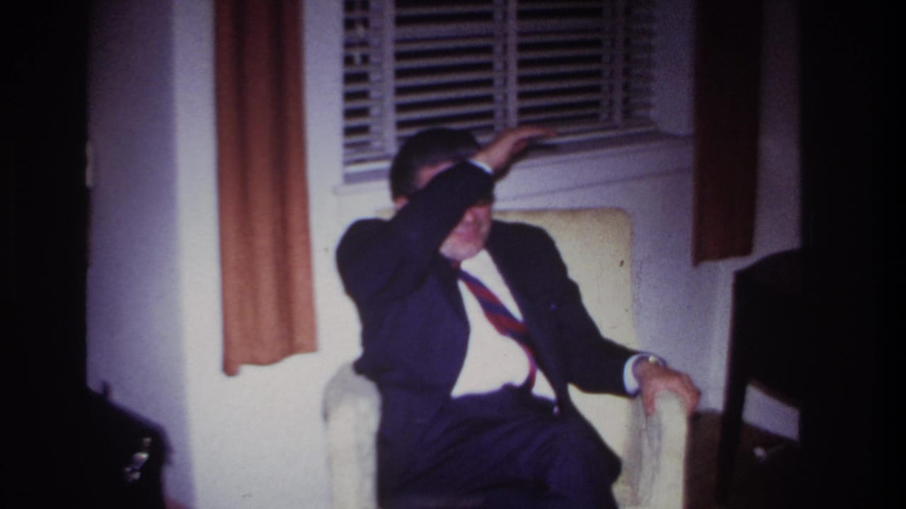 Man in a suit sits in a chair while raising his arm toward his face.