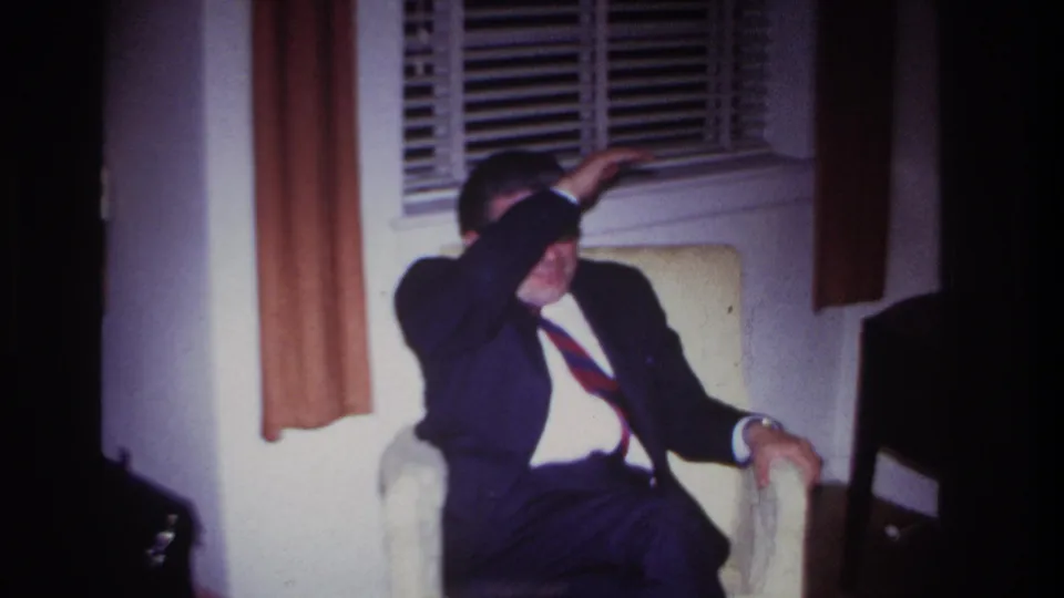 1967 Rockaway Beach interior frame with a suited man seated near venetian blinds, shielding his eyes from light.