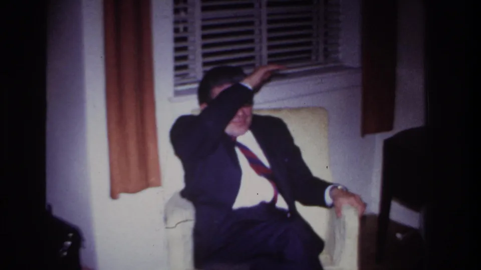 1967 Rockaway Beach home-movie frame of a suited man in an armchair reacting to bright light in a private room.