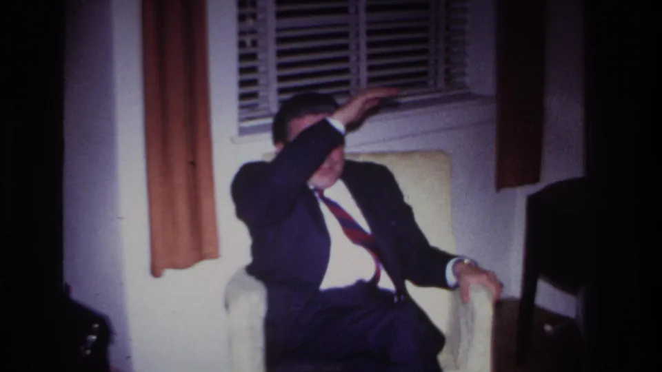 1967 Rockaway Beach interior frame showing a suited man seated by blinds with his arm raised against the light.