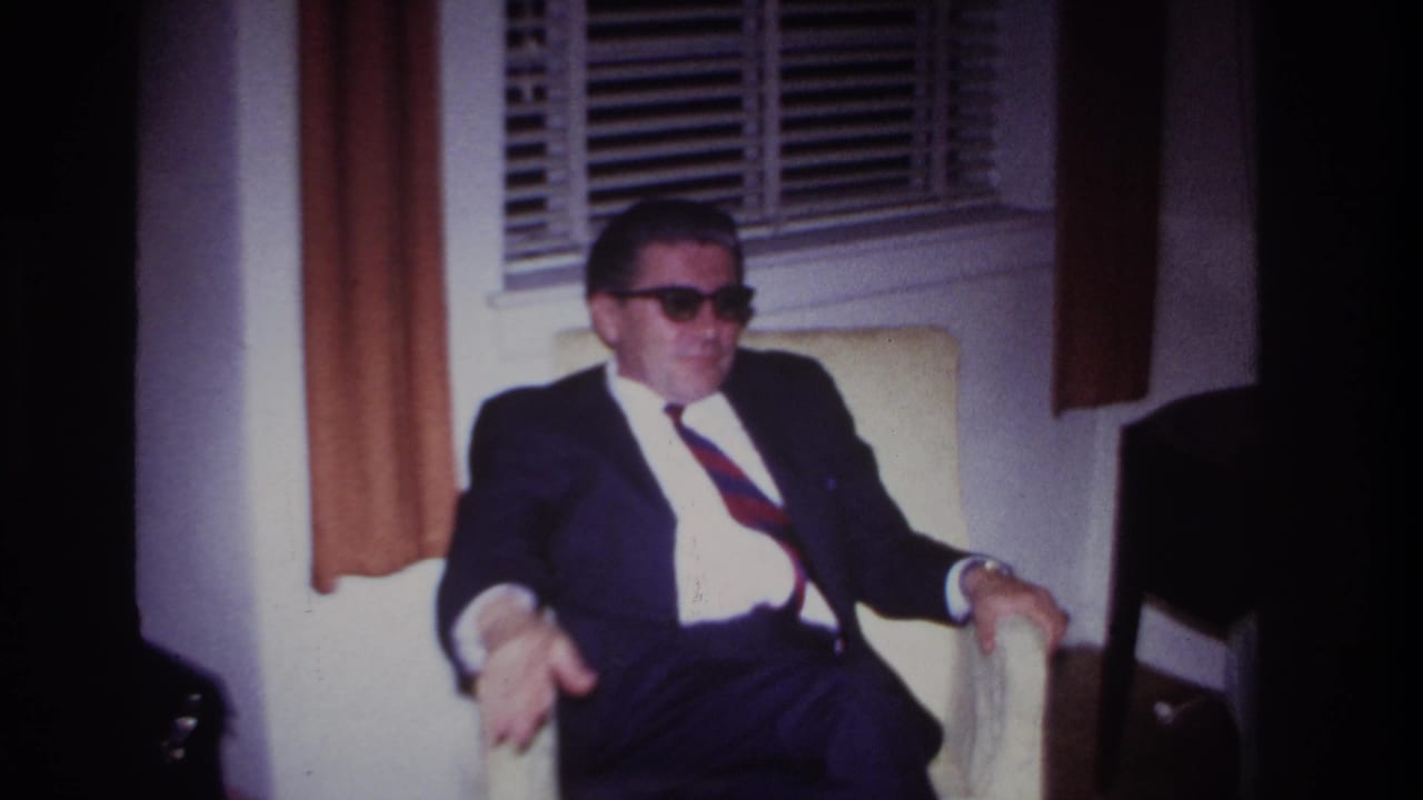 In a 1967 Rockaway Beach home-movie frame, a suited man with sunglasses sits indoors by venetian blinds and brown curtains.