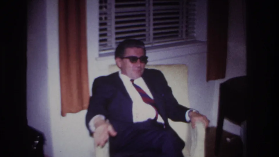 In a 1967 Rockaway Beach home-movie frame, a suited man with sunglasses sits indoors by venetian blinds and brown curtains.