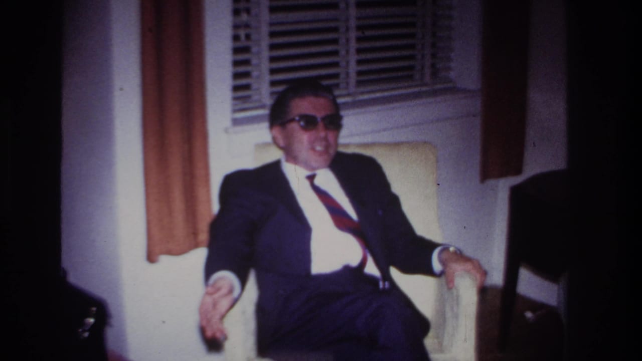 The seated man in sunglasses gestures with one hand while sitting in a Rockaway Beach interior, with blinds and curtains behind him.