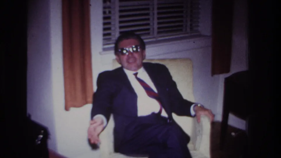 A 1960s home-movie frame shows the suited man leaning in the armchair, sunglasses reflecting light as he gestures indoors.