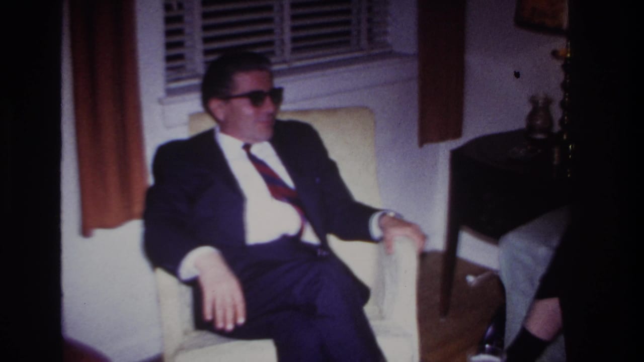 In a 1967 Rockaway Beach home-movie frame, a suited man wearing sunglasses sits in a cream armchair beside window blinds and curtains.