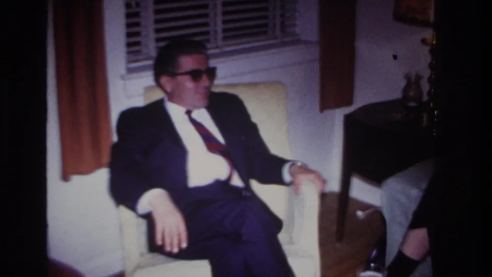 In a 1967 Rockaway Beach home-movie frame, a suited man wearing sunglasses sits in a cream armchair beside window blinds and curtains.