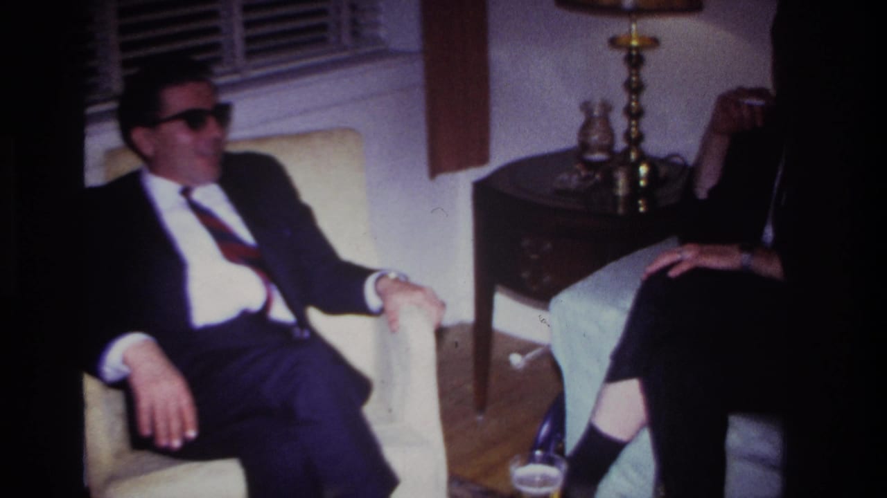 Man in sunglasses seated beside another adult partly visible in a living room.