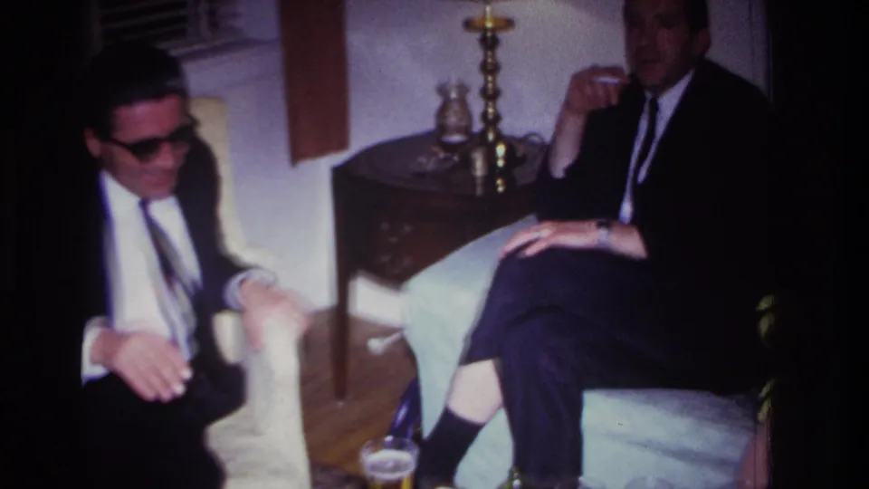 Two formally dressed adults sit near upholstered chairs, a wooden side table, brass lamp, and glassware in the 1967 home-movie interior.