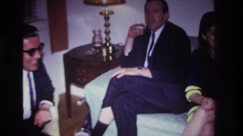 A suited man and a woman in a dark dress with yellow trim sit with others in the Rockaway Beach private living-room gathering.