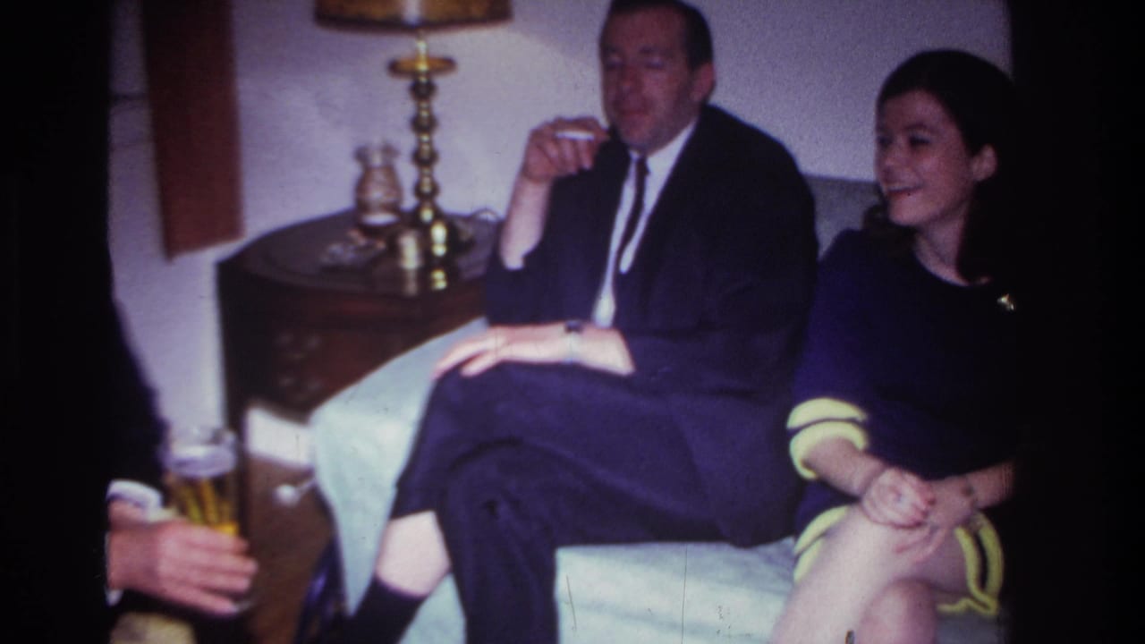Man in a dark suit smoking on a couch beside a young woman in a dark dress, with another person holding beer at left.