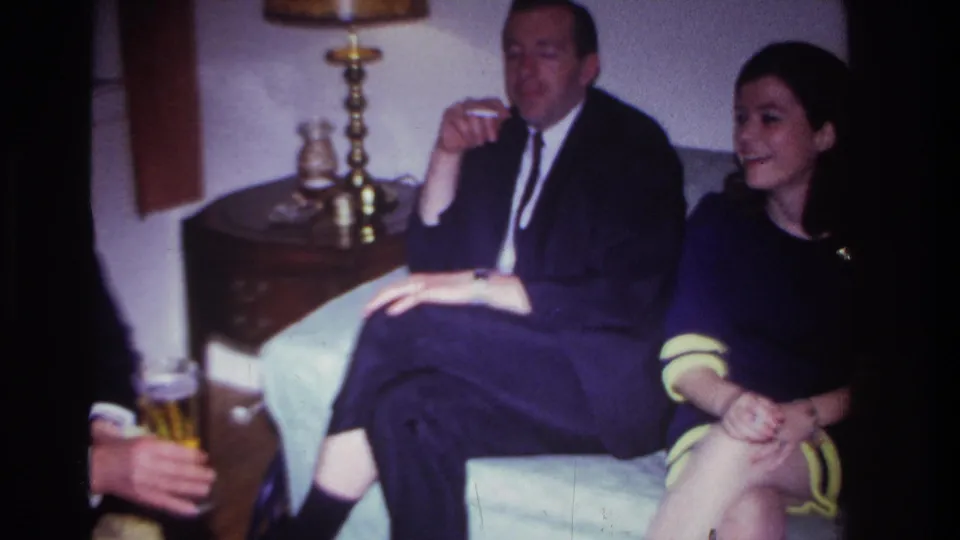 1967 Rockaway Beach home-movie frame showing a suited man smoking on a couch beside a smiling young woman in a dark dress with yellow trim.