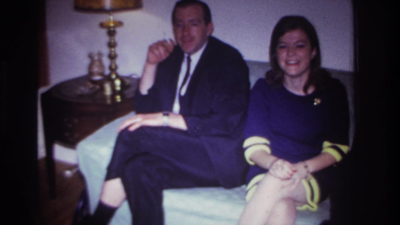 1967 Rockaway Beach interior frame with a suited man and smiling young woman seated closely on a light couch in a private living room.