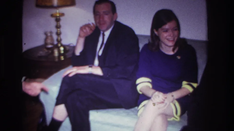 1967 Rockaway Beach home-movie frame showing a smiling young woman seated beside a man in a suit in a private living-room gathering.