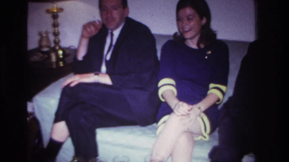 1967 Rockaway Beach frame of a woman in a dark dress with yellow trim sitting beside a suited man on a couch during a family gathering.