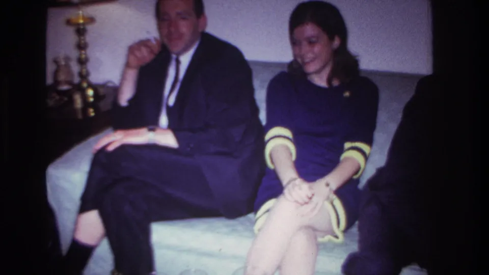 1967 Rockaway Beach home-movie frame showing a suited man with a cigarette and a woman in a purple dress seated on a couch.