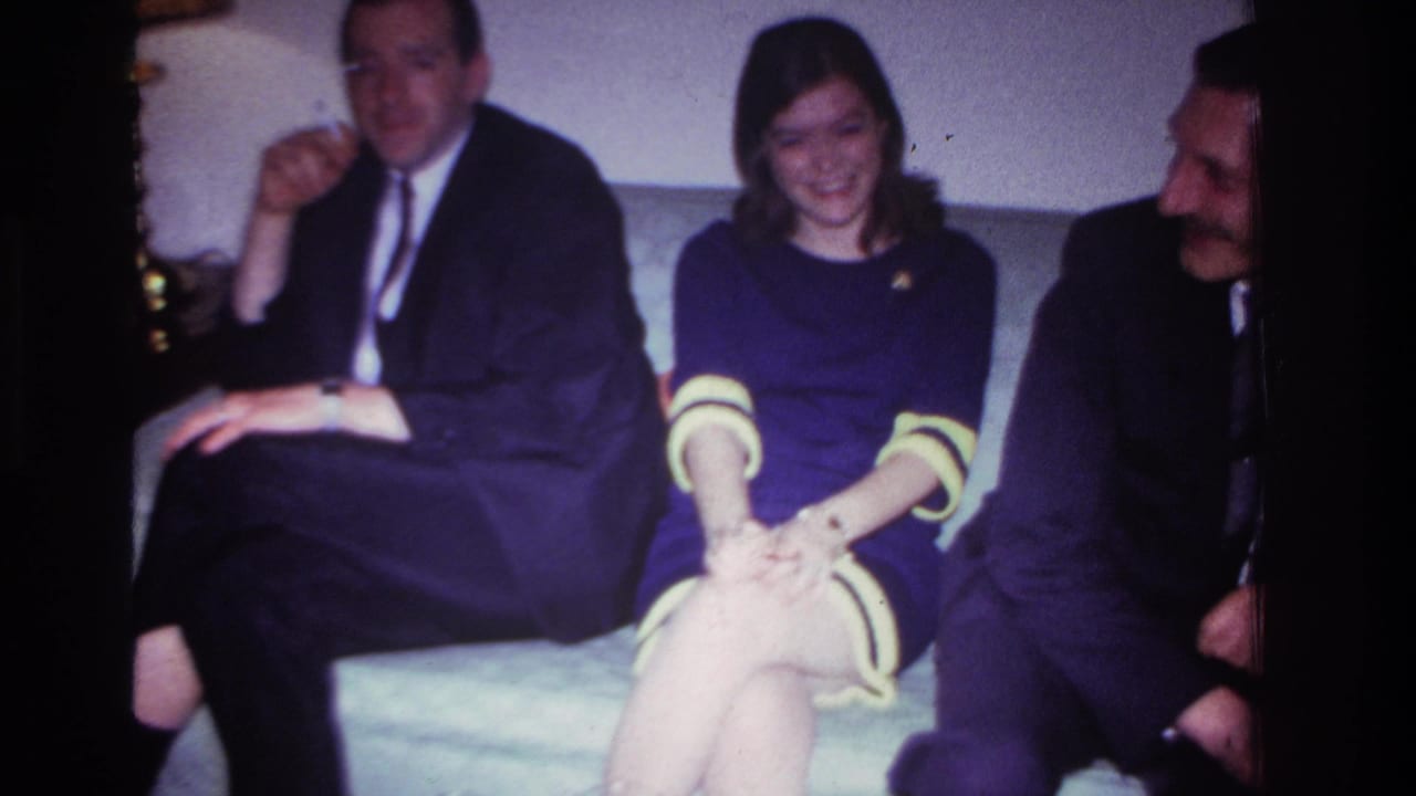 1967 Rockaway Beach interior frame with three adults on a couch, the woman smiling while the men sit on either side.