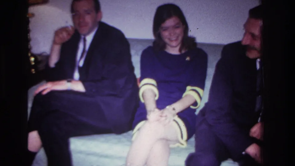 1967 Rockaway Beach interior frame with three adults on a couch, the woman smiling while the men sit on either side.