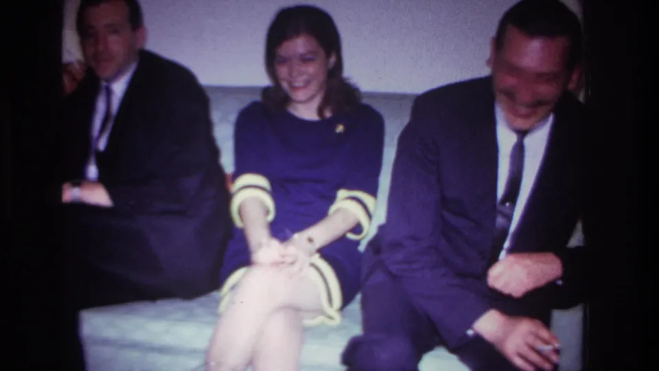 1967 Rockaway Beach home-movie frame showing a smiling woman seated between two suited men in a living room.