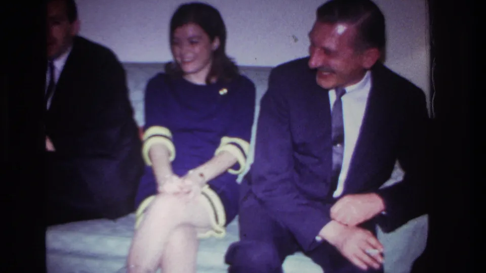 1967 Rockaway Beach living-room frame with a woman smiling toward a suited man seated close beside her.