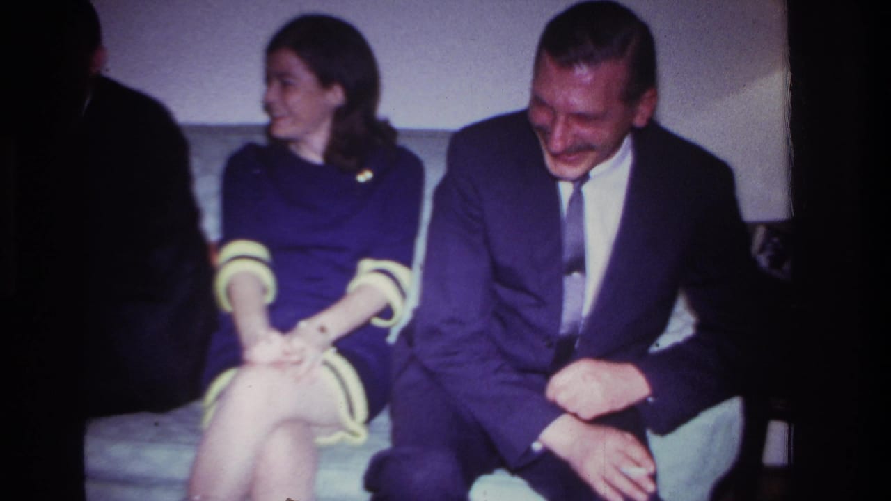 A suited man laughs on a couch beside a smiling woman in a purple dress.