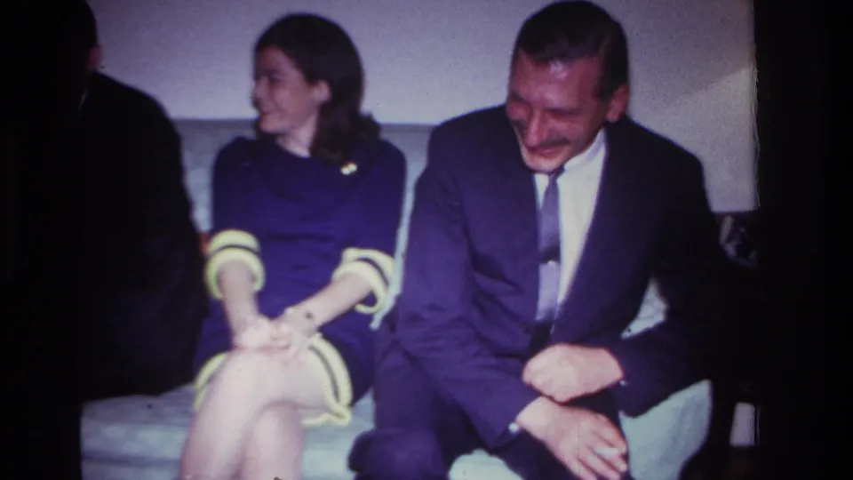 1967 Rockaway Beach home-movie frame showing a suited man laughing beside a woman in a purple dress on a living-room couch.