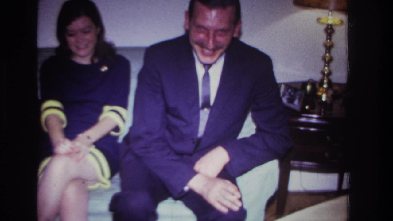 1967 Rockaway Beach living-room frame with a woman in a purple dress and a suited man seated near a wooden table and lamp.