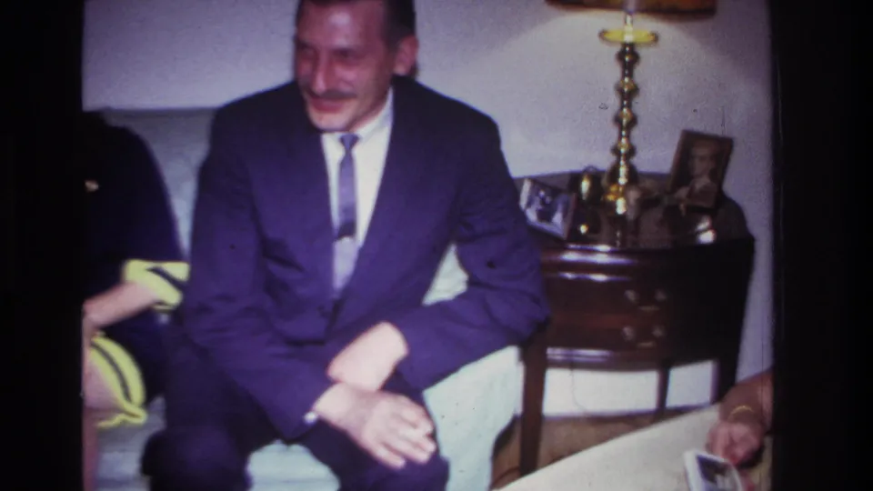 1967 Rockaway Beach home interior frame showing a suited man on a couch beside a lamp, side table, and framed photos.
