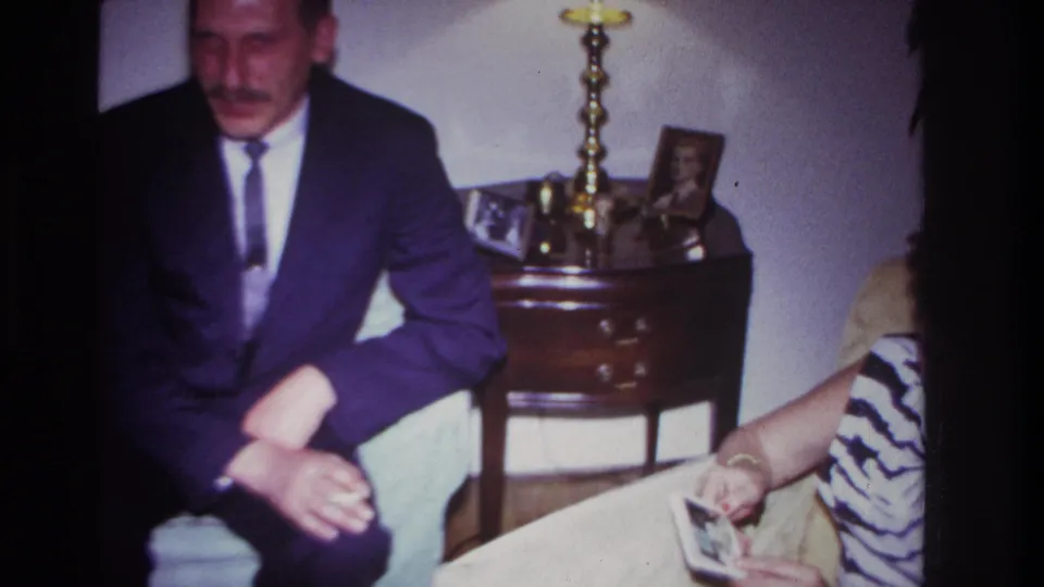 1967 Rockaway Beach frame with a suited man on a couch, lamp table behind him, and a partially visible guest holding photos.