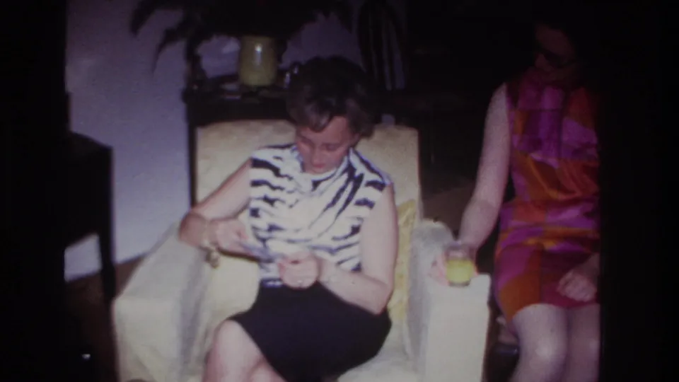 1967 home-movie still from Rockaway Beach showing two women seated indoors, one handling photographs and the other holding a drink.