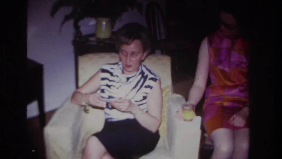 Rockaway Beach 1967 frame of a seated woman looking up from photographs, with another woman in a colorful dress beside her in a private room.