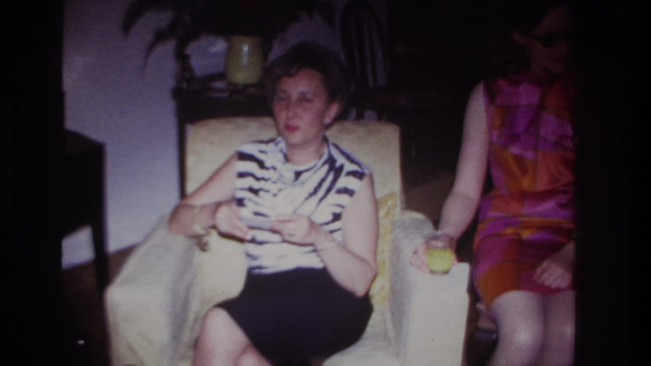 Two women seated indoors, one in a pale armchair holding a small object and another beside her with a drink.
