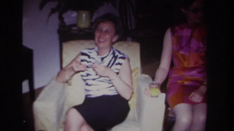 1967 Rockaway Beach frame showing a smiling seated woman in a living room beside a woman in sunglasses and a bright dress.