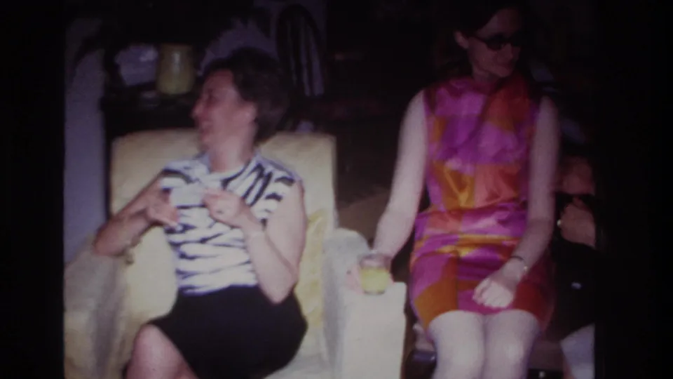 1967 Rockaway Beach home-movie frame of two women laughing and seated close together inside a private living room.