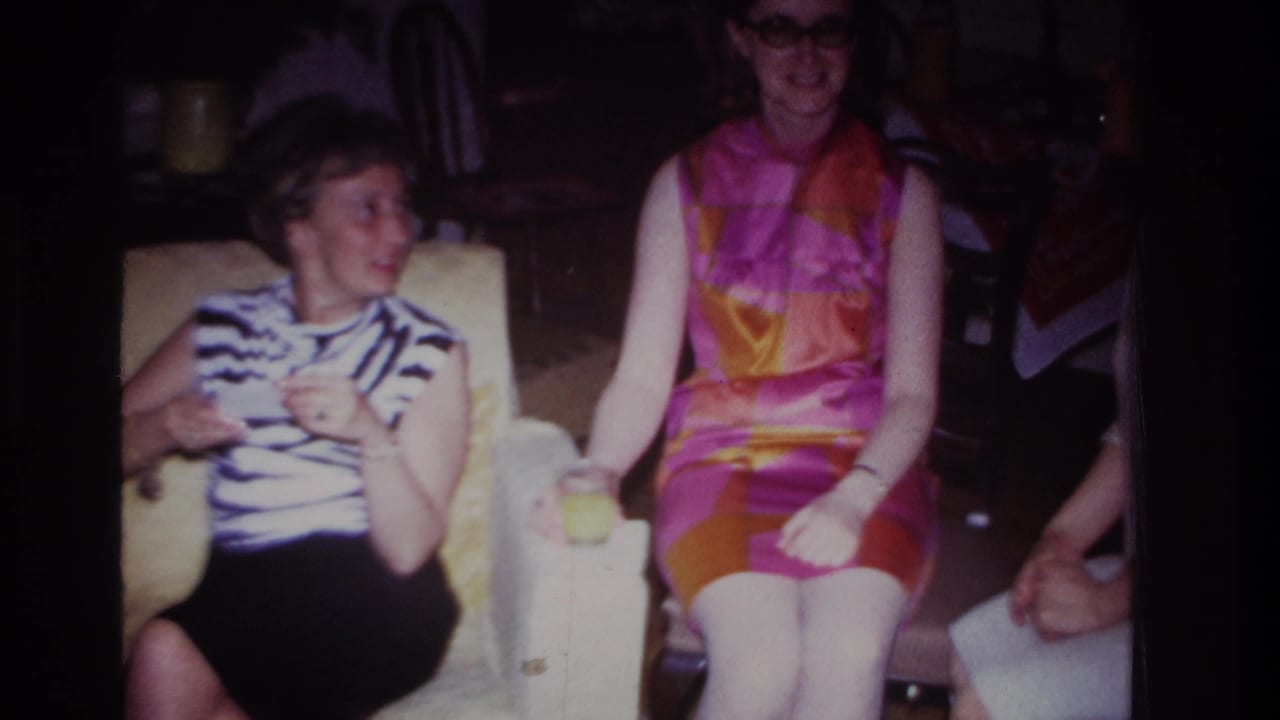 Two women in 1960s attire sit indoors with drinks, one turning toward someone off frame while the other faces forward.