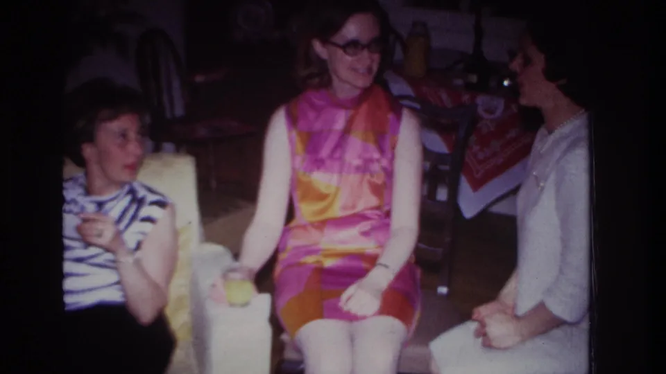 Three women in a 1967 home interior converse beside a couch and yellow chair, with drinks visible and a patterned cloth in the background.