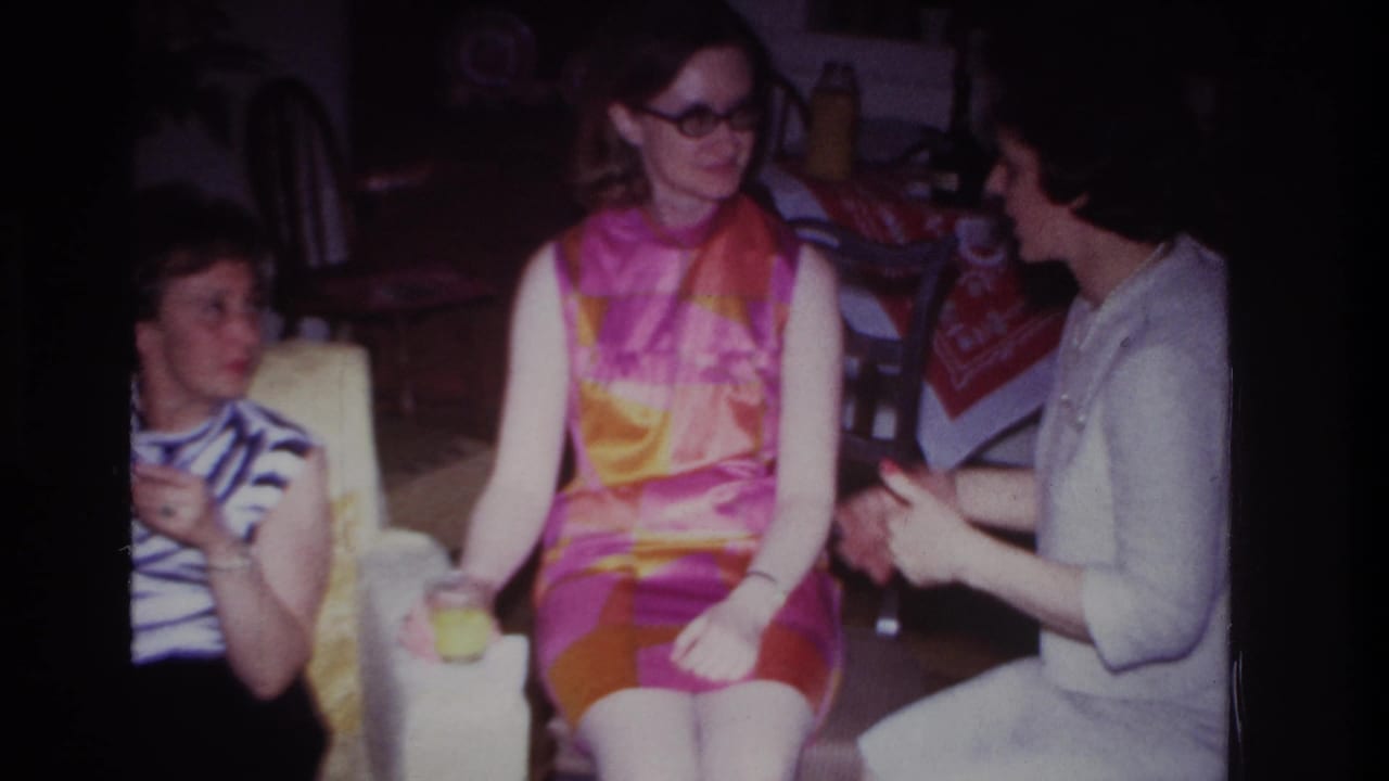 1967 Rockaway Beach home-movie frame showing three women seated indoors, with the central woman in glasses and a pink-orange dress.