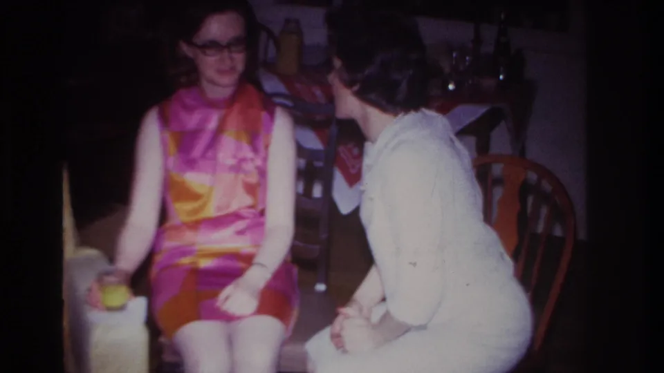 1967 Rockaway Beach home interior frame showing two seated women in conversation, one in a vivid patterned dress and glasses.